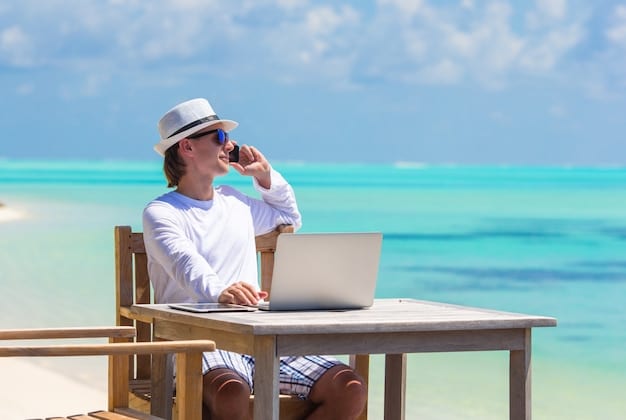 A person happily working remotely from a beach, laptop open, drinking a tropical beverage, with a clear blue sea and palm trees in the background.